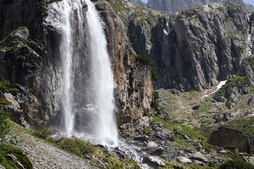 A tall waterfall dropping down a rock face in an alpine valley under a clear sky