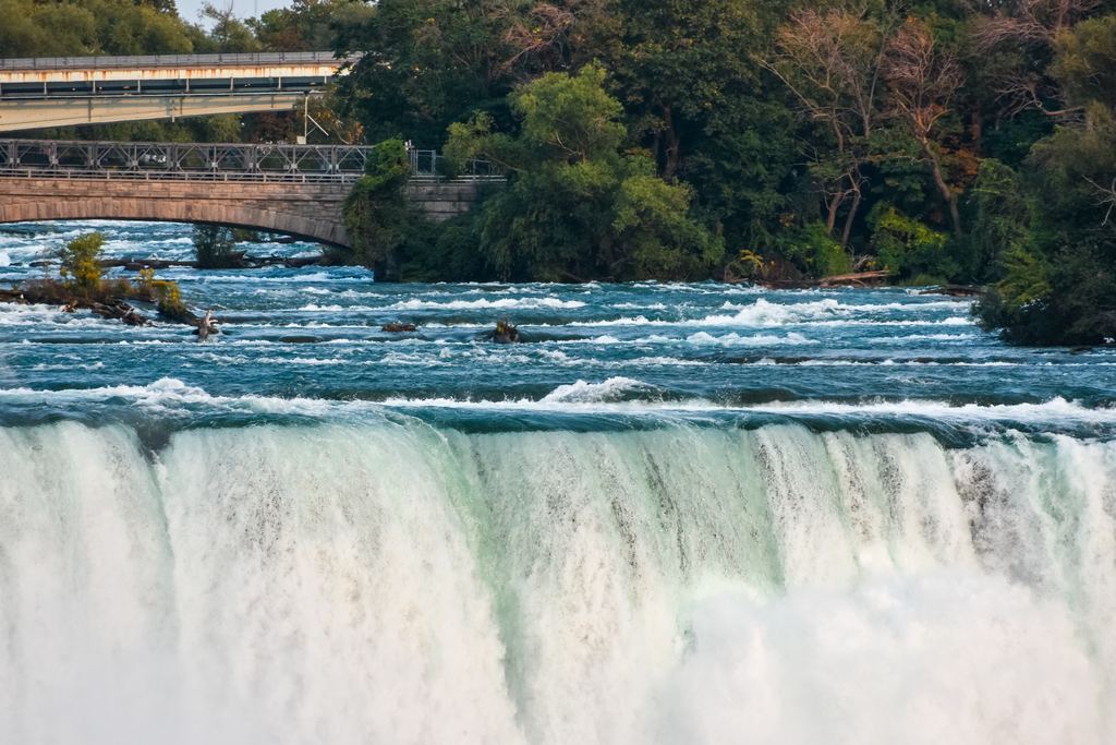 The crest of the American Falls at Niagara, with river rapids above the edge and a bridge in the background