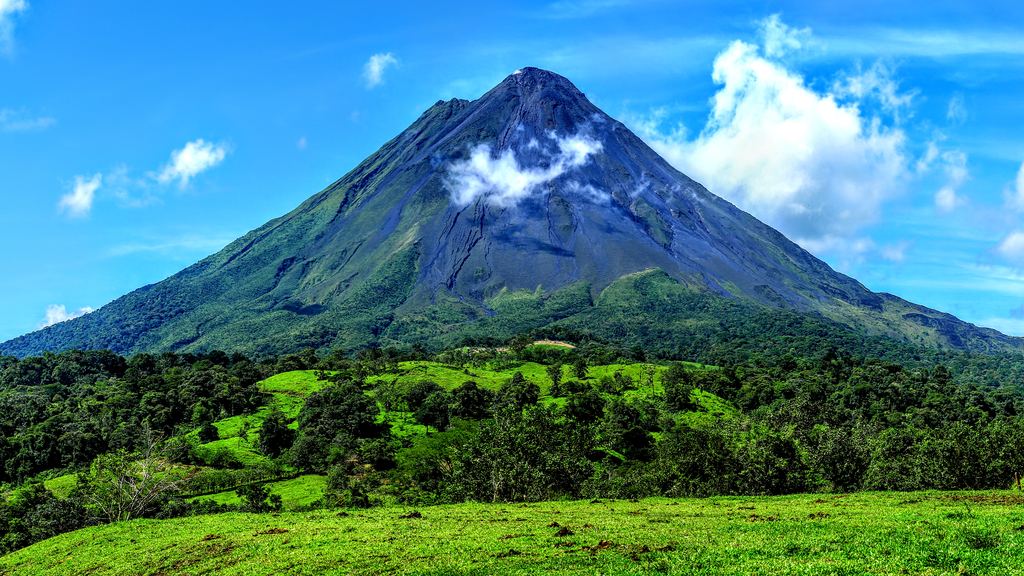 Arenal volcano under a blue sky with a cloud on its slope, above forest and green pasture in Costa Rica