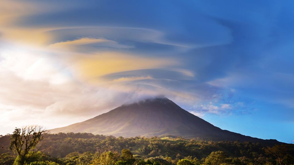 Arenal volcano rises above green pasture and forest under a sky of clouds at sunset, Costa Rica