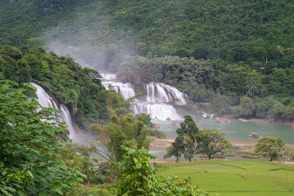 Ban Gioc Falls seen across green fields from a viewpoint, with boats on the river in Cao Bang