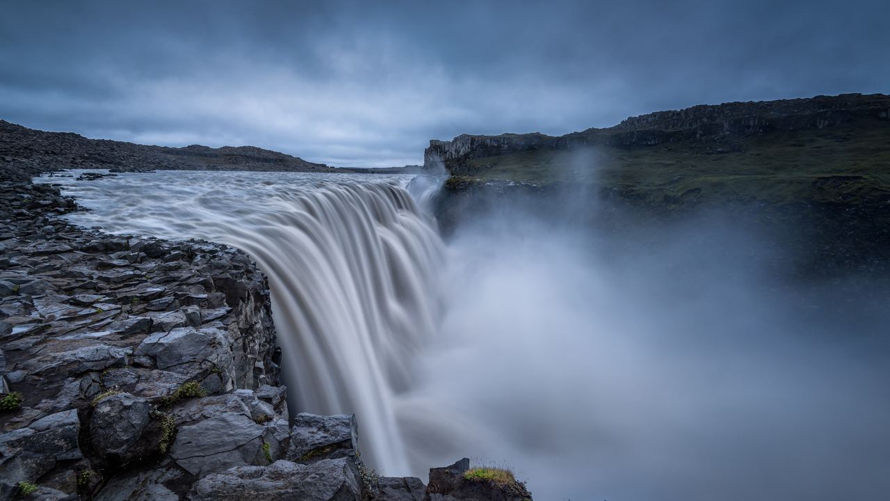 A broad waterfall dropping over a rocky rim under a dark cloudy sky with mist rising