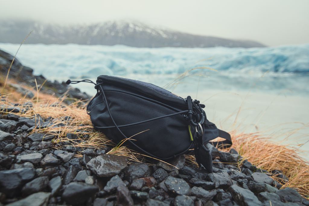 A black camera bag on dark gravel beside a glacial lake with mountains in the haze behind
