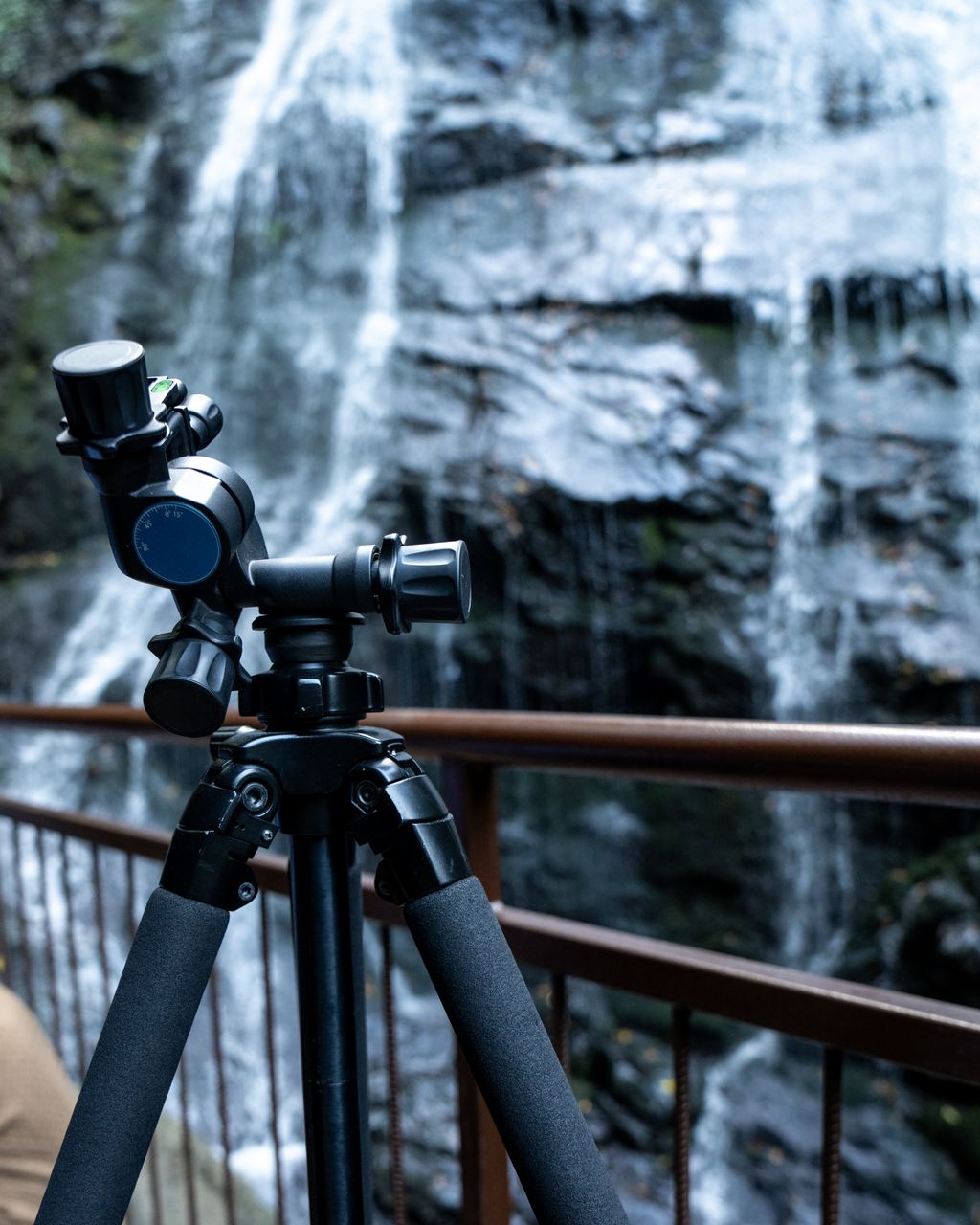 A camera tripod set up at a rusty railing in front of a waterfall running down a dark cliff