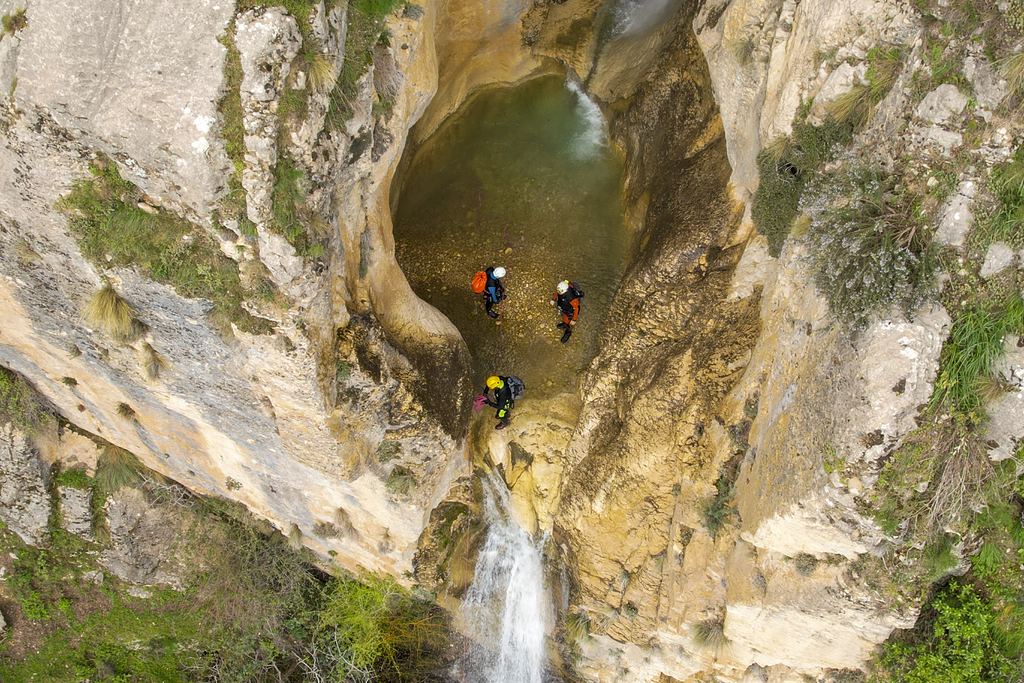 Aerial view of three canyoners at a pool between rock walls below a waterfall