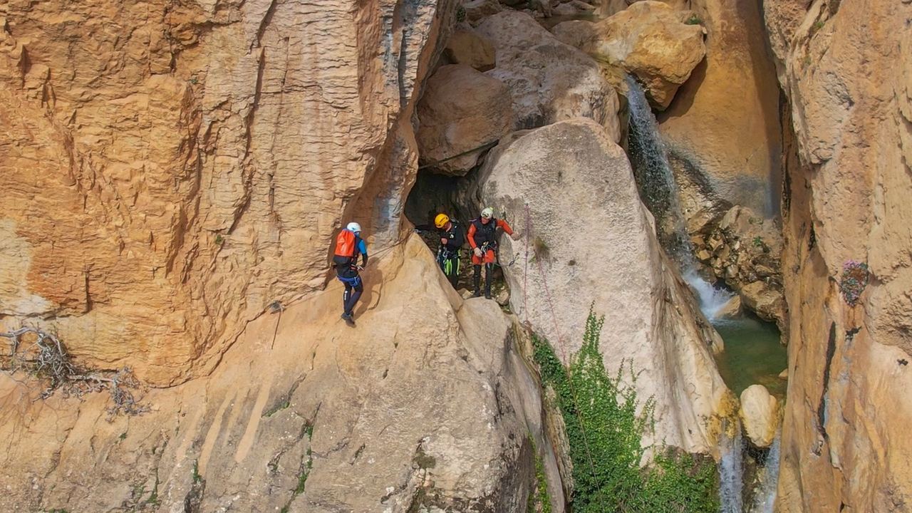 Three canyoners with helmets and packs on a ledge in an orange rock gorge beside a waterfall