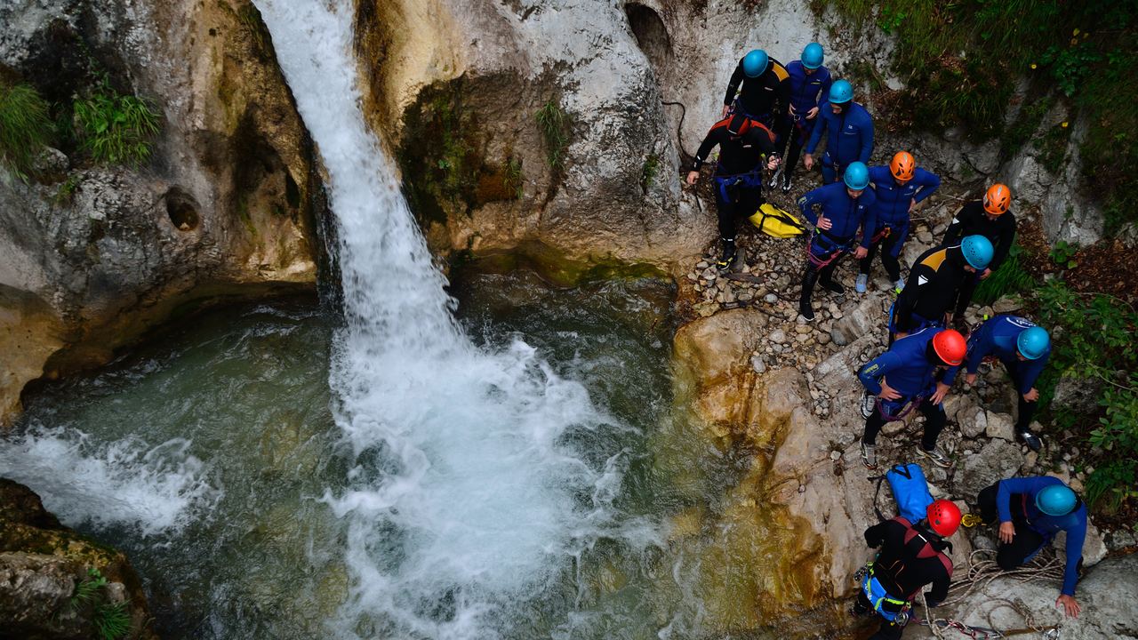 Group in helmets and wetsuits gathered at a pool below a waterfall in a rocky canyon
