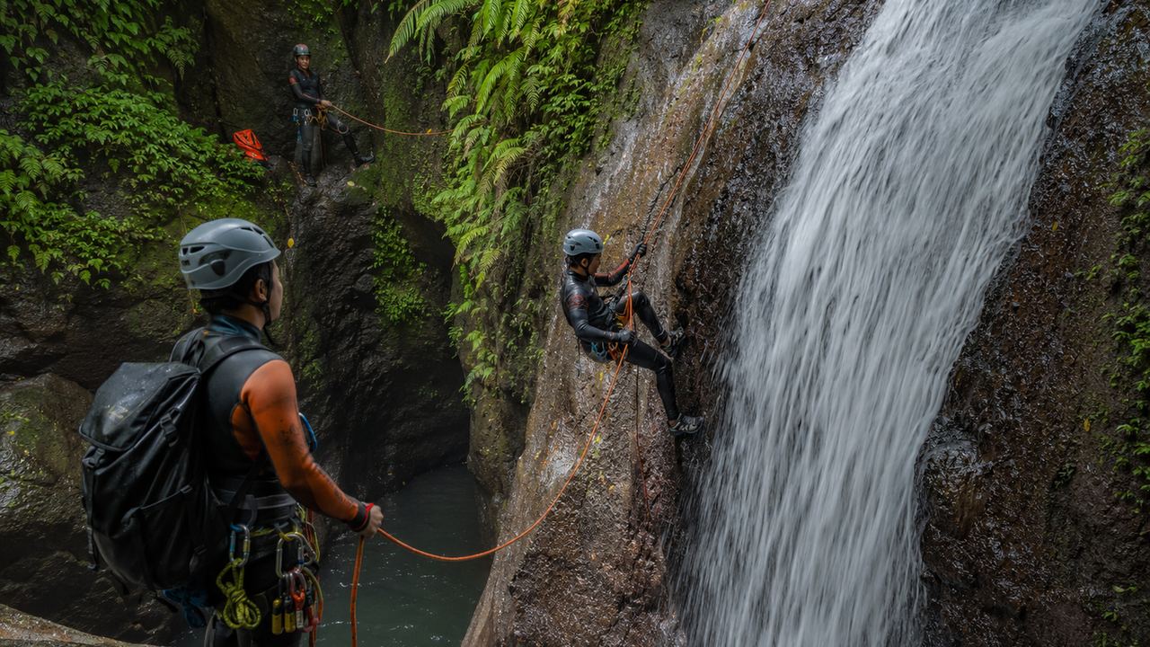 Two canyoning guides with helmets and ropes at the foot of a tall waterfall in a fern-lined canyon