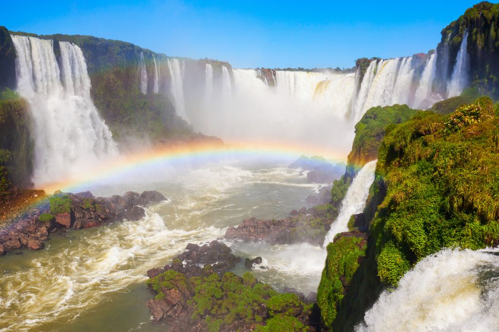 Rainbow over the river below the Devil's Throat at Iguazu Falls with moss-covered rocks