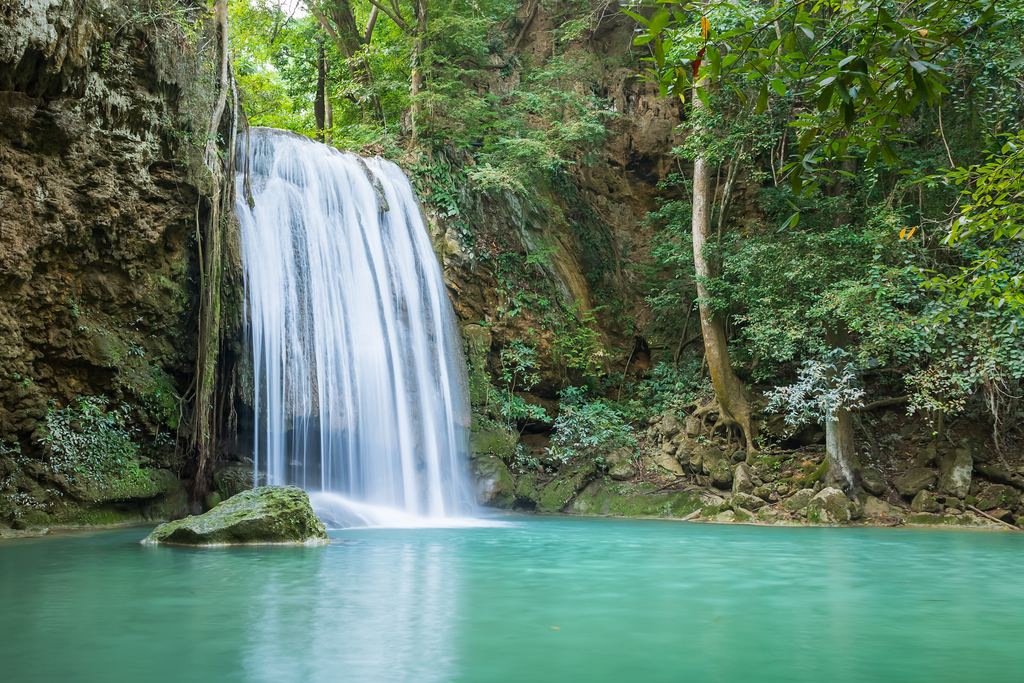 A waterfall pouring into a green pool in a forest at Erawan Falls, Thailand