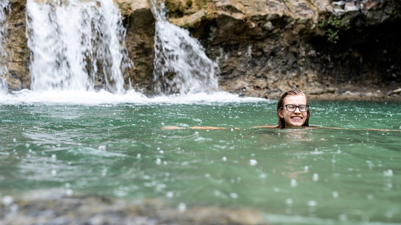 Swimmer in a green pool below a small waterfall in a rocky forest