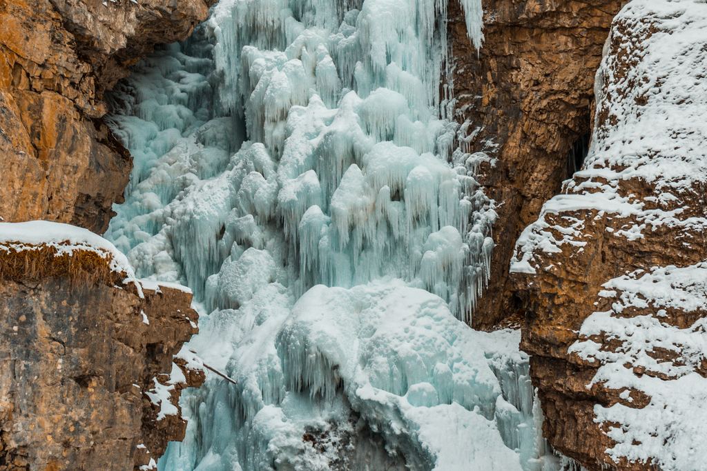 A tall frozen waterfall hanging between rock walls above a frozen pool, with snow on the ledges