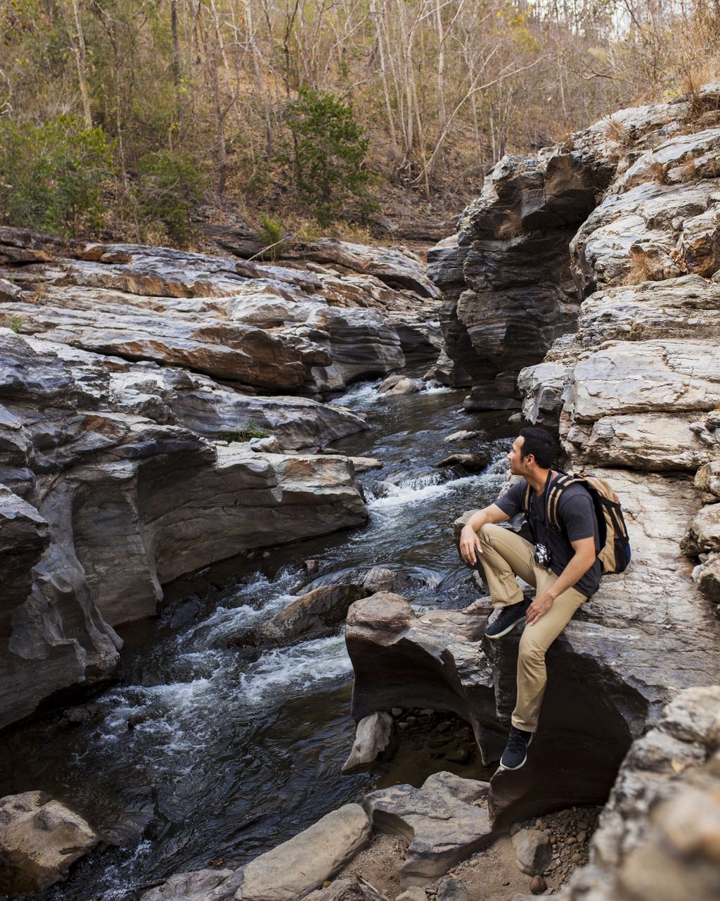A hiker with a backpack resting on rocks beside a stream in a rocky gorge