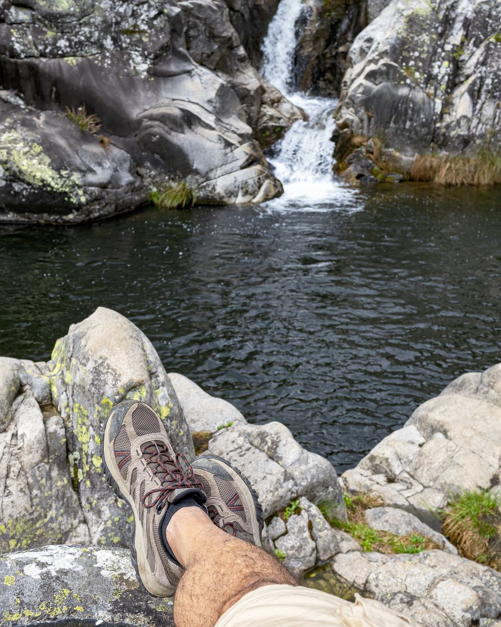 Hiking shoes crossed on lichen-covered granite above a dark pool with a small waterfall