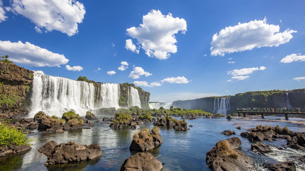 Wide view of the Iguazu Falls from a viewpoint with a catwalk over rocks and a blue sky