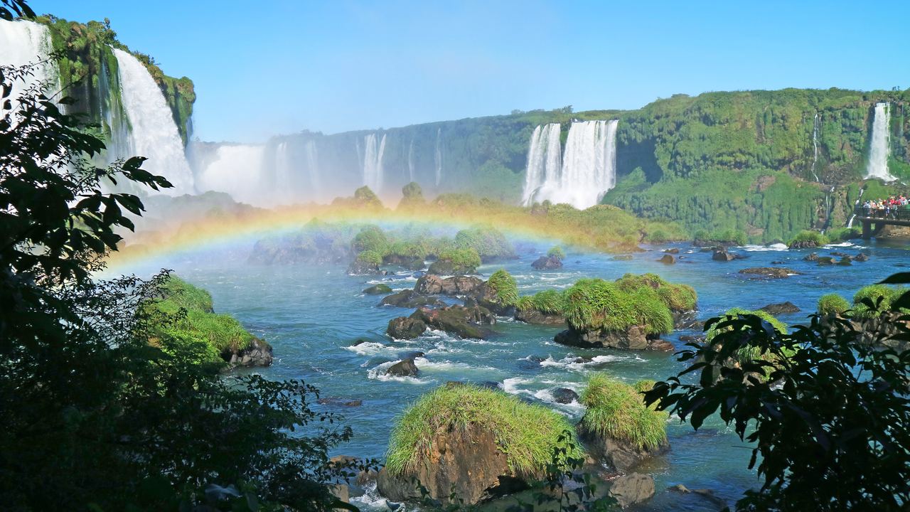 The curtain of Iguazu Falls seen from the Brazilian side, with a rainbow over the river and rainforest on both banks