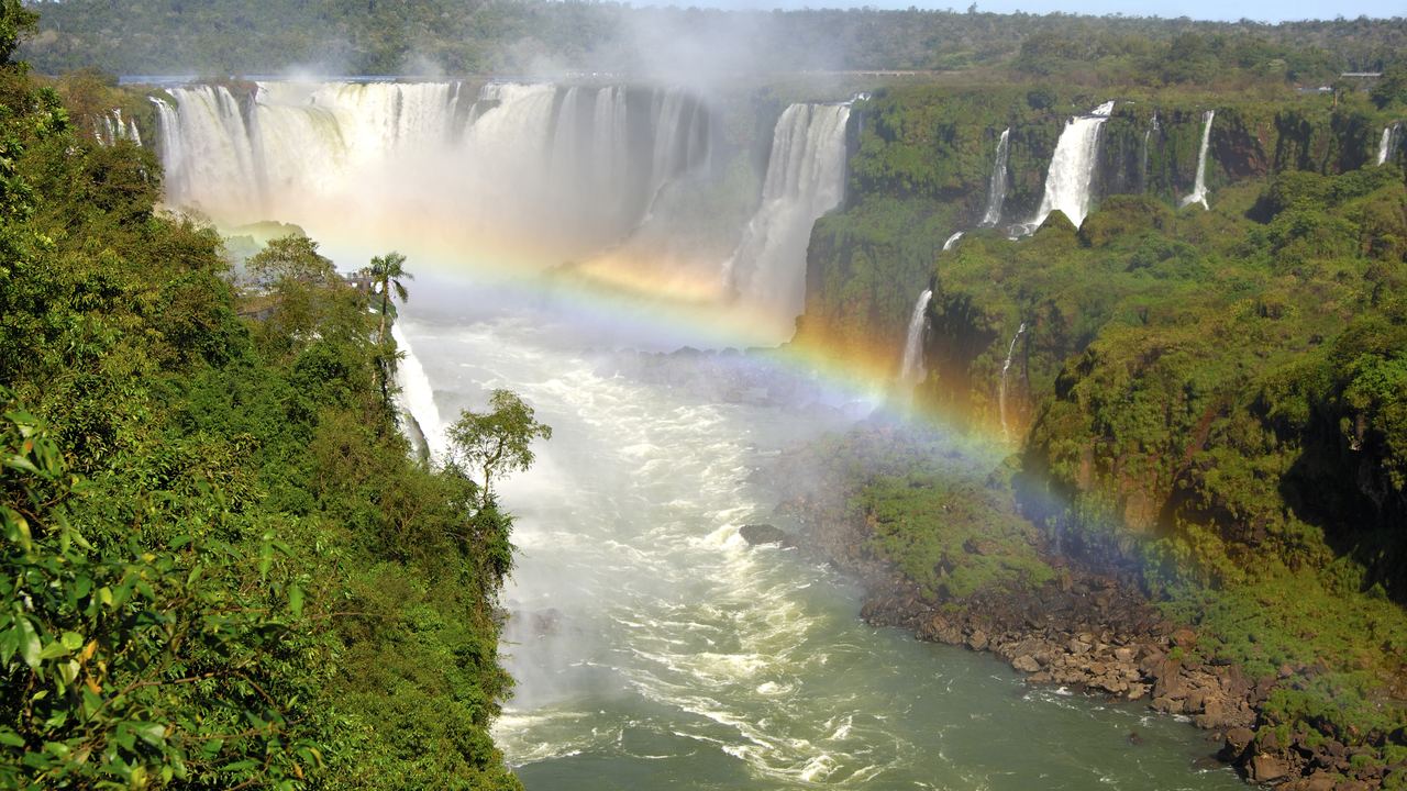 Wide view of the Iguazu Falls with a rainbow over the river and forest on both banks