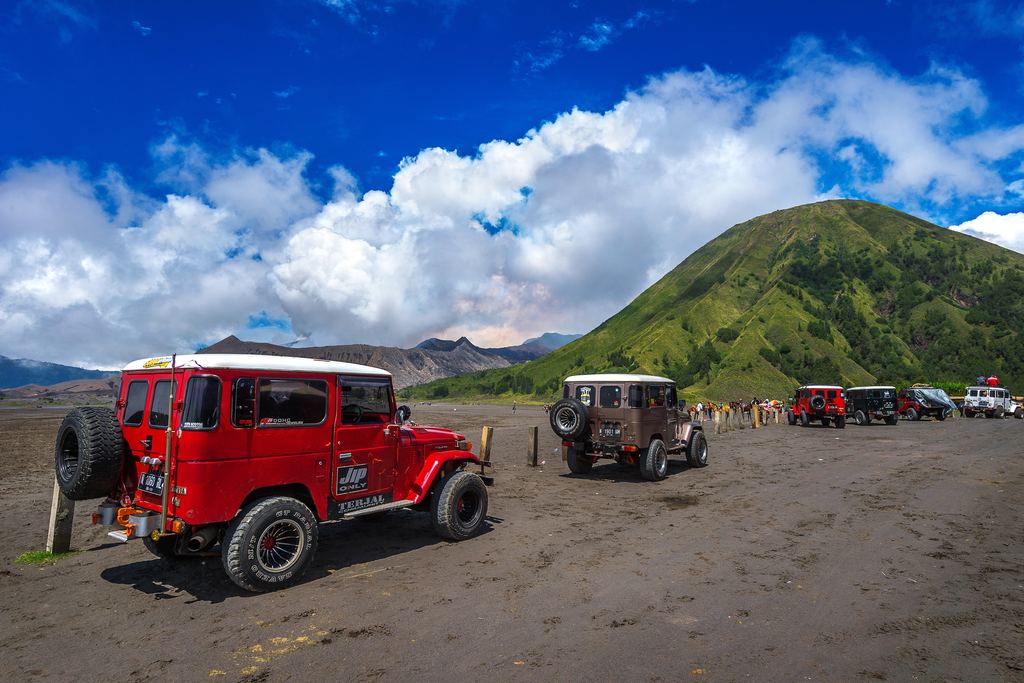 Red and brown 4WD jeeps parked on the dark volcanic sand at Mount Bromo below a green hill