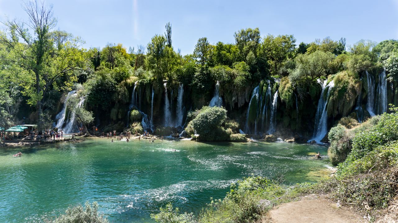 Wide view of the Kravica Waterfalls and the turquoise lake with visitors on the shore
