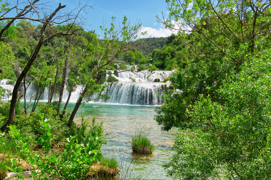 A wide waterfall over a tufa barrier into a turquoise pool, seen through trees in Krka National Park, Croatia