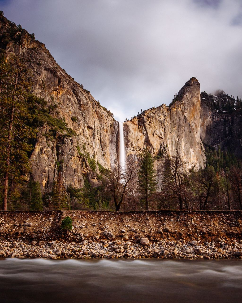 Long-exposure photo of a thin waterfall dropping from a granite cliff above a river