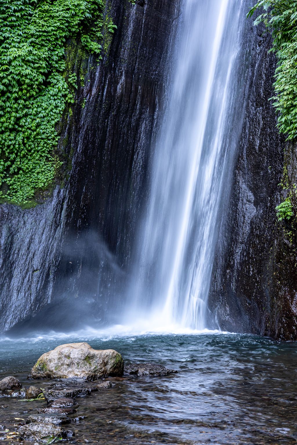 Narrow waterfall falling over a dark cliff into a rocky pool at Munduk in North Bali