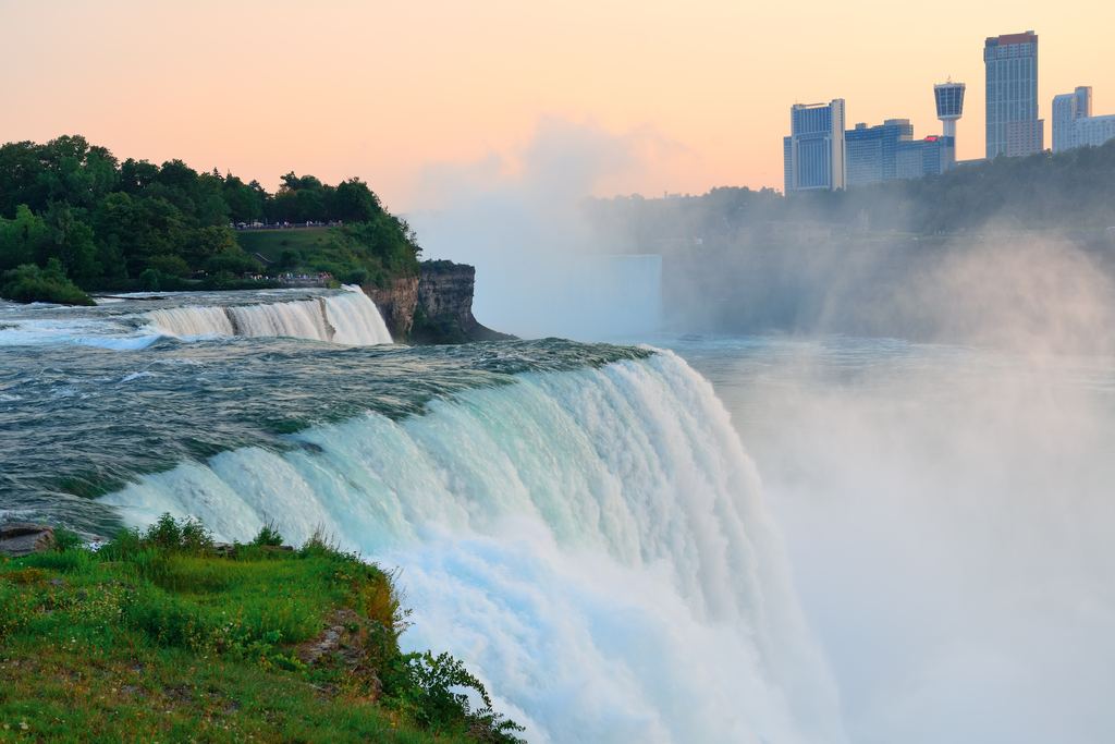 Niagara Falls at dusk with mist rising off the crest and the city skyline in the distance