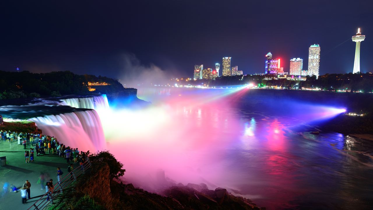 Niagara Falls lit in pink and blue at night, with visitors at a railing and hotel towers behind