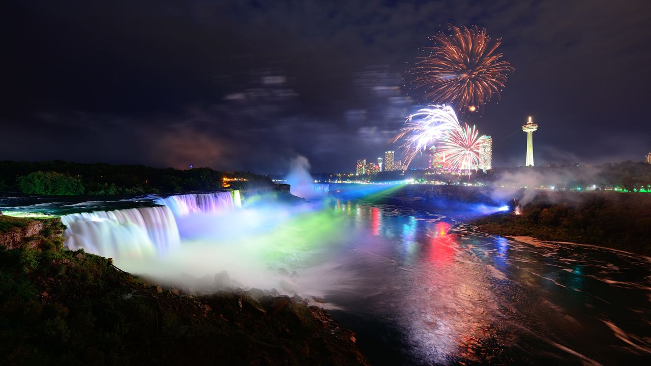 American Falls lit in green and white at night with fireworks above Niagara Falls and the Skylon Tower