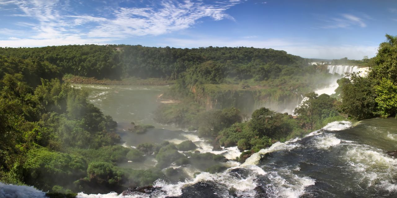 Wide panorama of Iguazu Falls and the rainforest around them, with white water rushing in the foreground