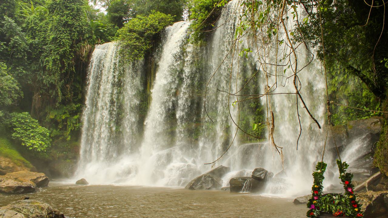 Wide Phnom Kulen waterfall falling into a brown pool, with a flower-decorated swing in the foreground