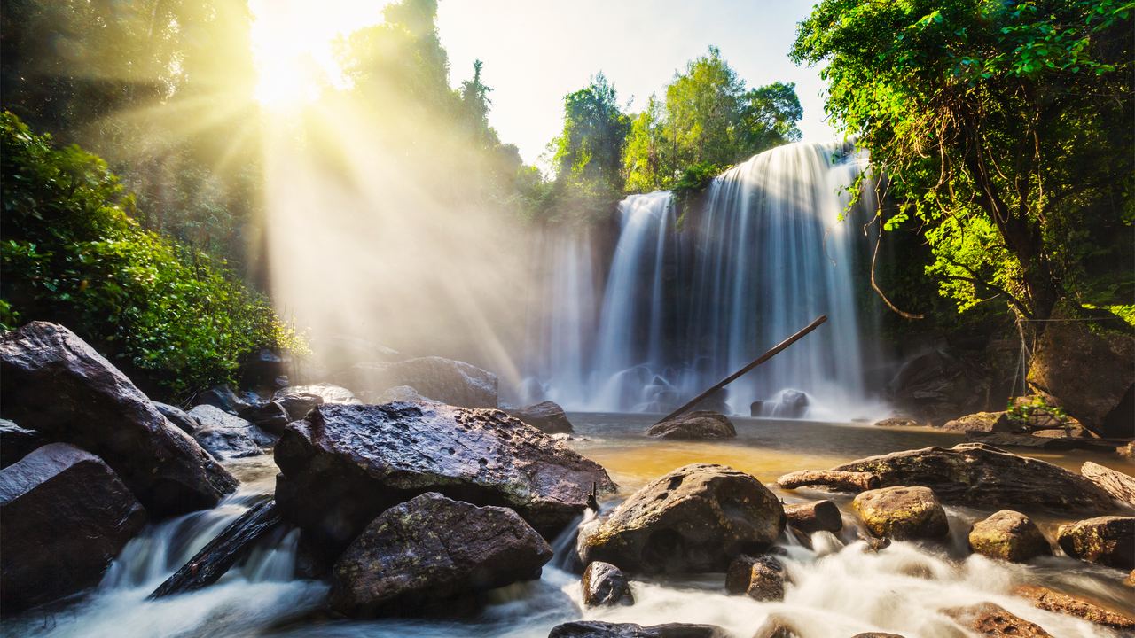 Phnom Kulen waterfall seen across rocks and a stream with sun rays through the trees