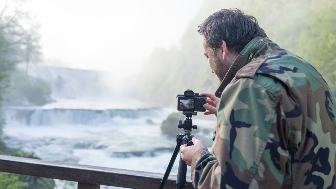 A photographer with a camera on a tripod at a wooden railing above white water and mist