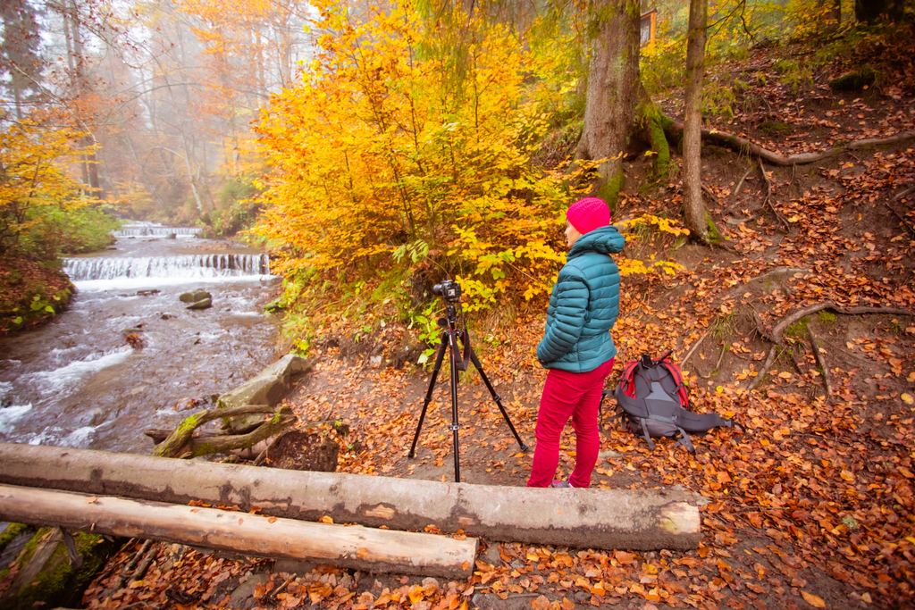 A photographer with a tripod and a backpack beside a stream with small falls in an autumn forest