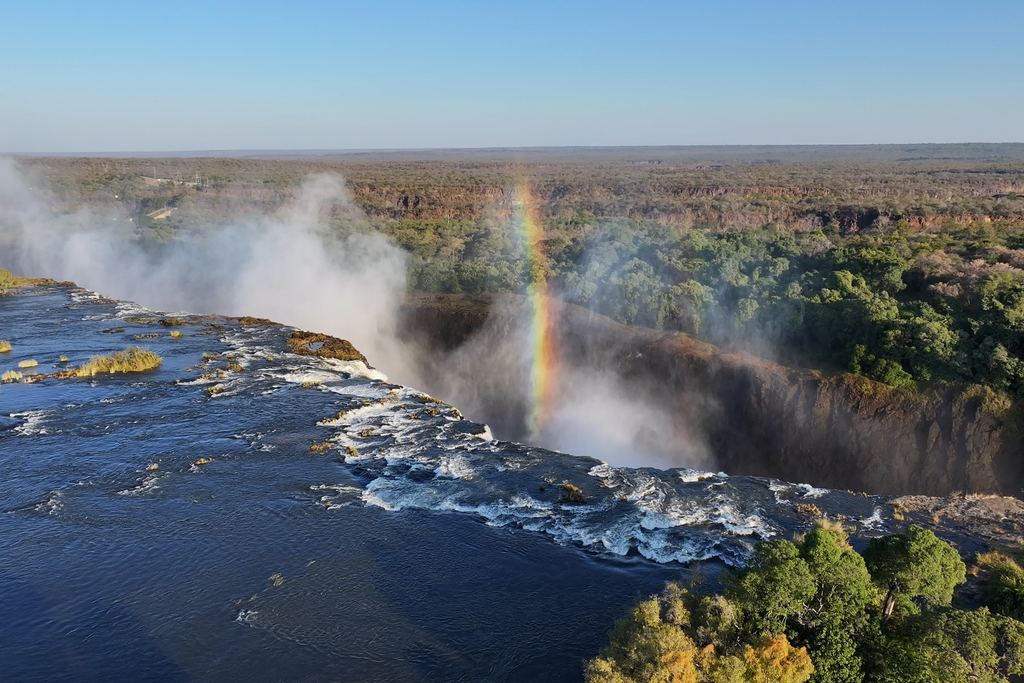 River running over the lip of Victoria Falls toward the gorge, with a rainbow standing in the spray