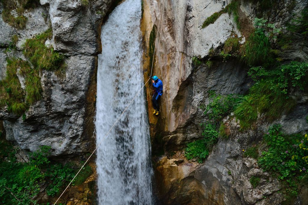 A canyoner wearing a helmet descending a rope beside a waterfall in a narrow rocky gorge