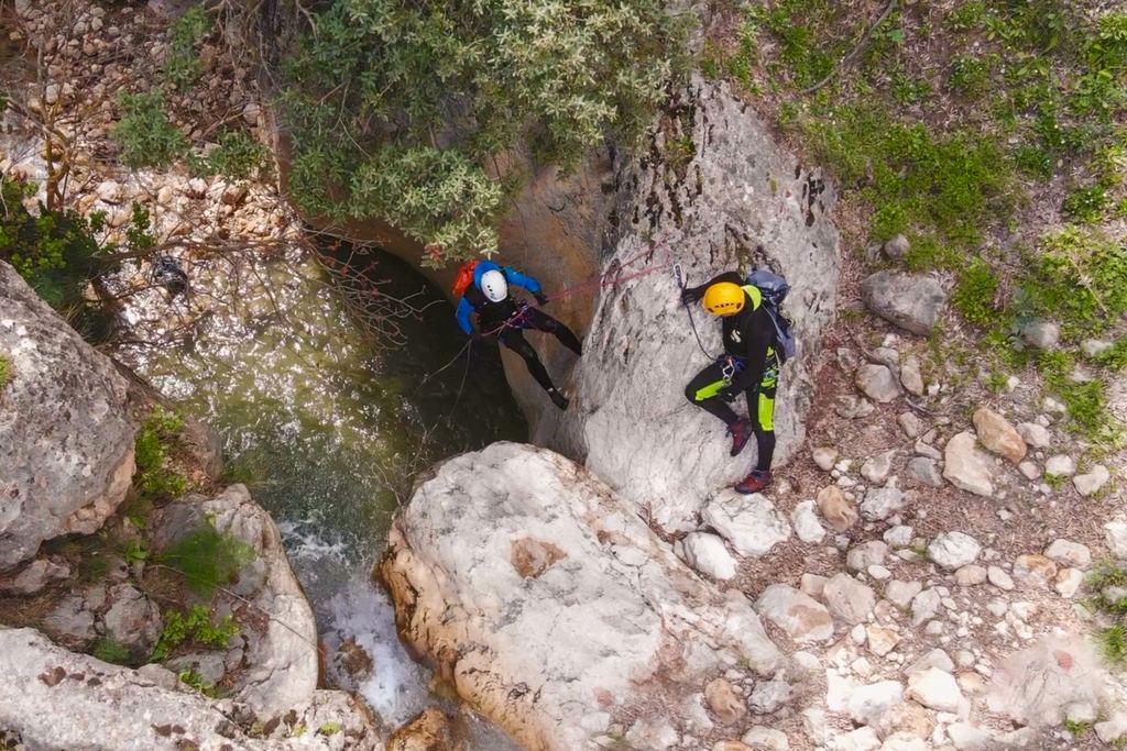 Two canyoners in helmets at a rappel station above a narrow pool in a rocky gorge