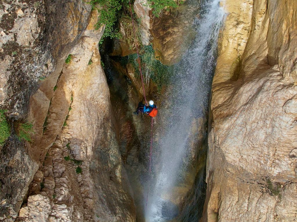 Seen from above, a canyoner hangs on a rope in a narrow rock slot beside a thin waterfall