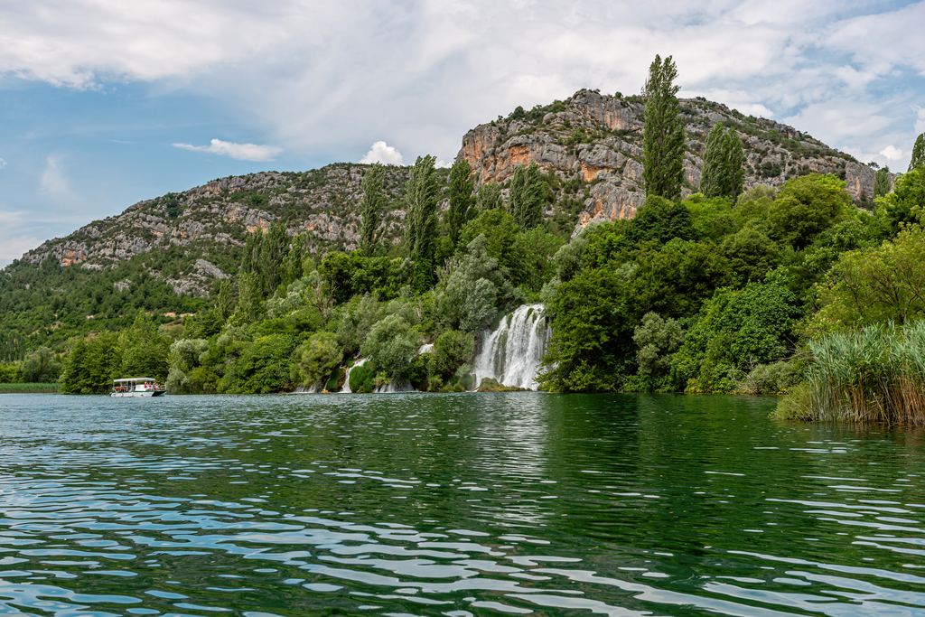 Roški slap falling from wooded cliffs into the Krka, with an excursion boat on the water, Krka National Park, Croatia