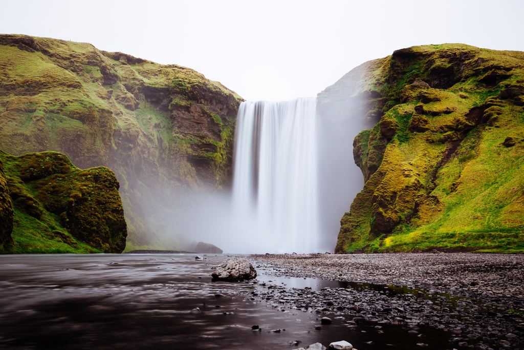 Skógafoss in long exposure, a 60 m curtain of water between two moss-covered cliffs, with a gravel flat in front