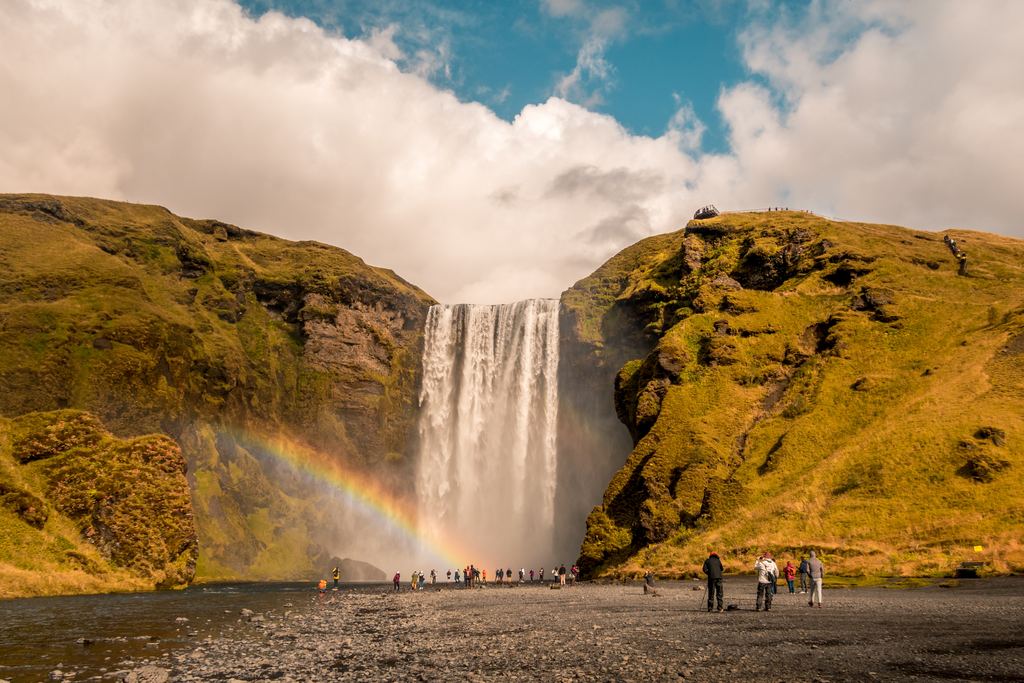 Skógafoss with a rainbow in the spray and visitors on the gravel in front