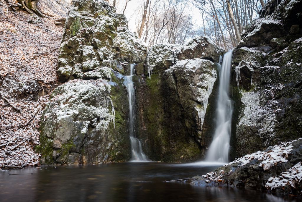 Two small waterfalls flowing between rocks lightly dusted with frost in a leafless winter forest