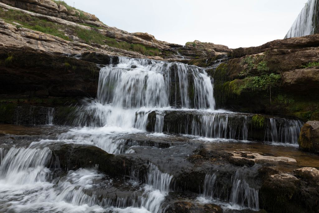 A stepped cascade flowing over moss-covered rock ledges under a pale sky