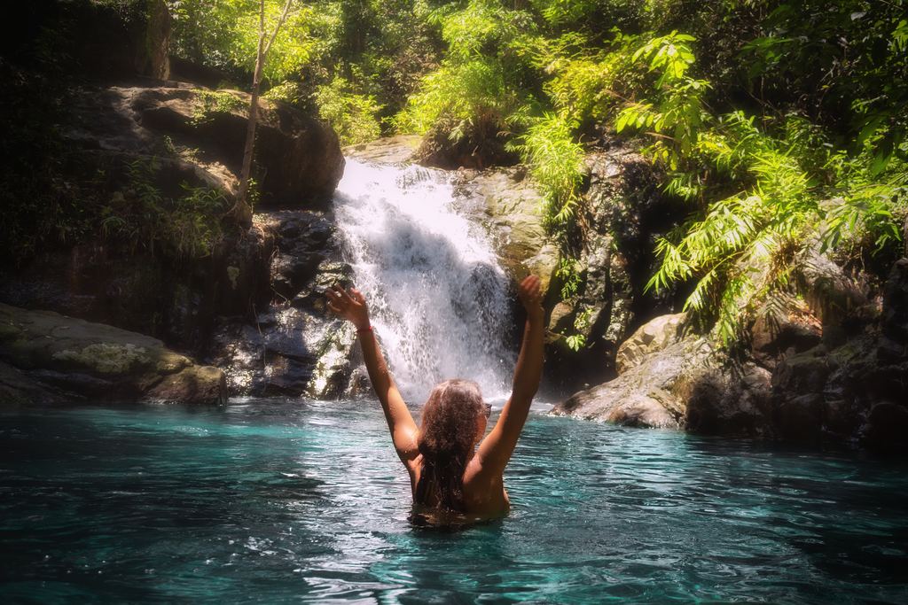 Swimmer with raised arms in a green pool below a small waterfall, with ferns and dark rock around