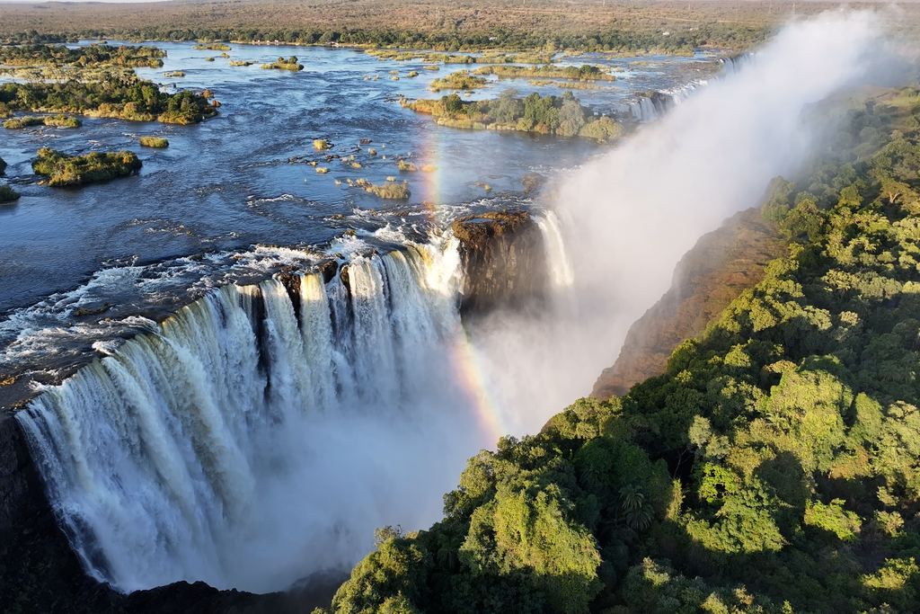 Aerial view of the Zambezi reaching the lip of Victoria Falls, with islands, a rainbow and the gorge forest
