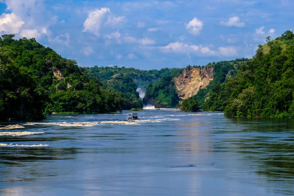 A tour boat on a wide river heading toward a waterfall in the distance between wooded hills
