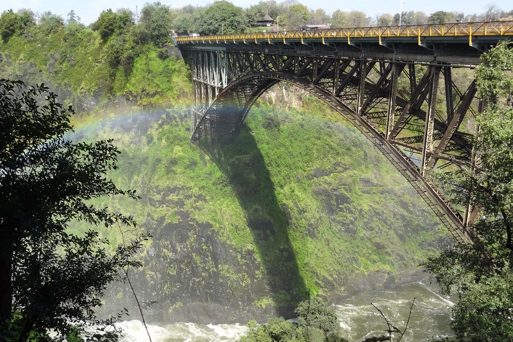 The steel arch of the Victoria Falls Bridge over the green gorge, with a rainbow forming in the spray below