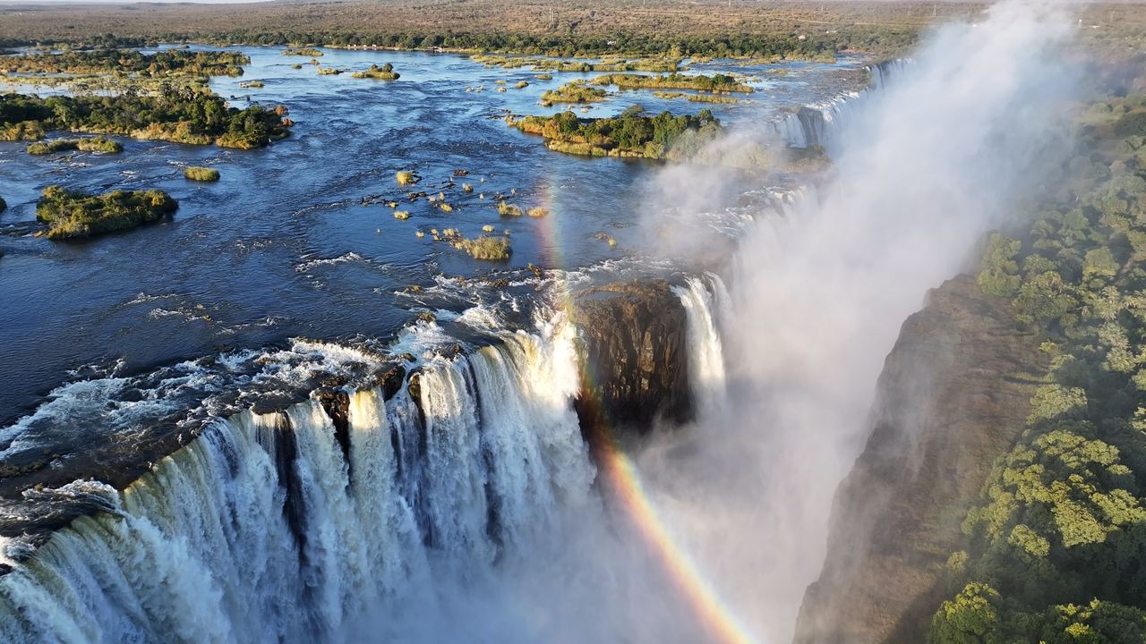 Aerial view of water pouring over Victoria Falls into the gorge, with a rainbow in the spray and forest beside it