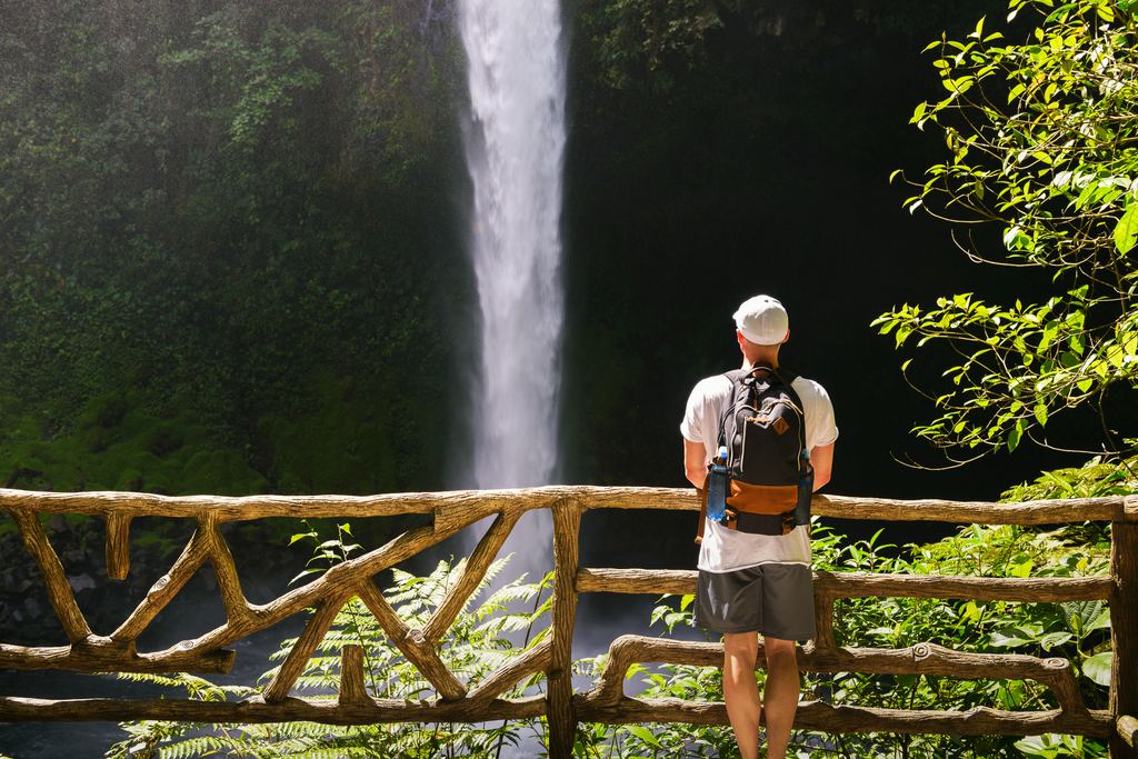 A hiker with a backpack stands at a wooden railing facing a tall waterfall in a Costa Rican forest