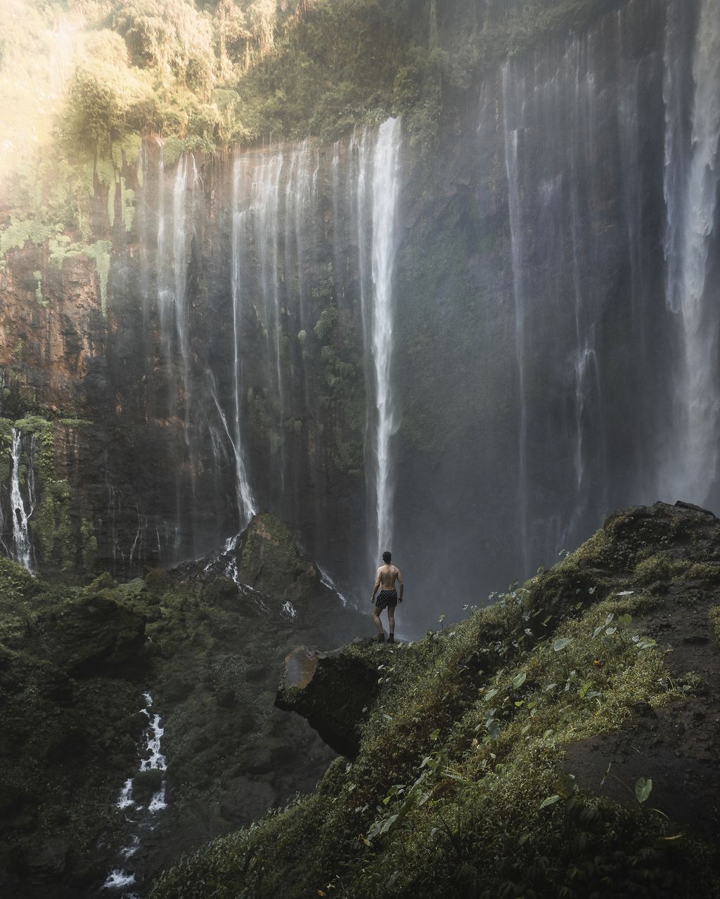 A hiker standing on a rock ledge in front of the wall of Tumpak Sewu waterfall
